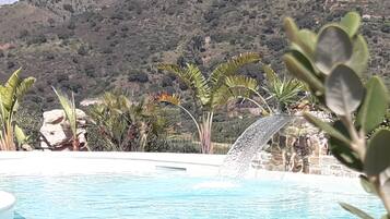 Une piscine agrémentée d’une cascade, entourée de plantes tropicales et bordée d’un paysage vallonné en arrière-plan.