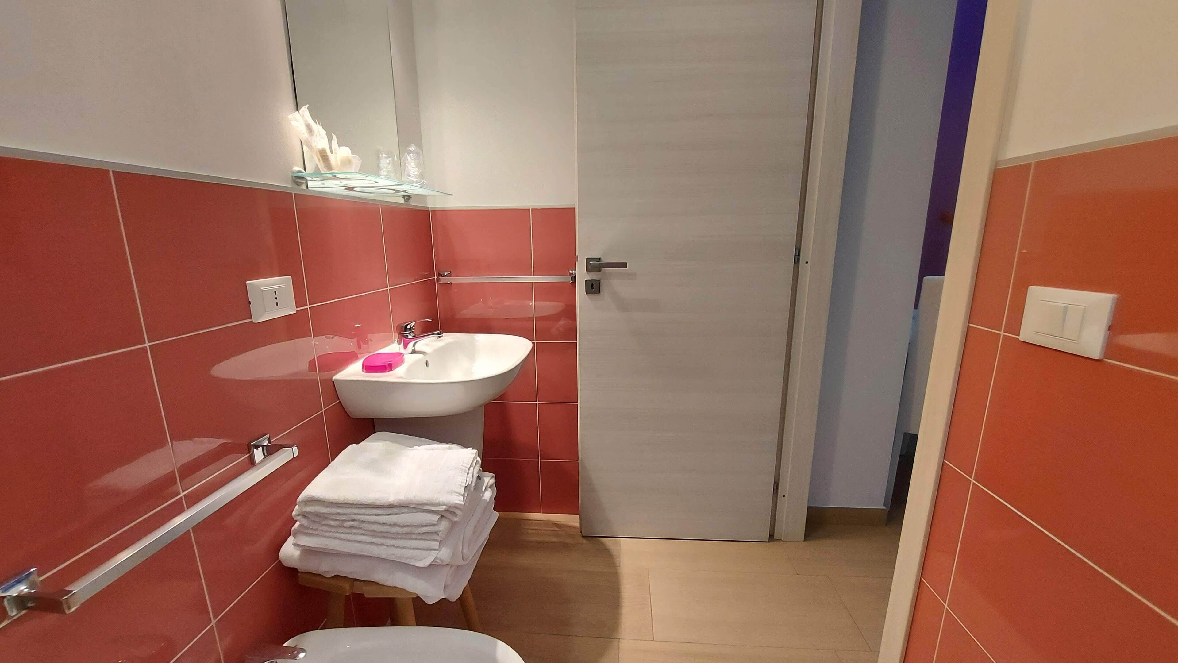 Un baño pequeño con un lavamanos blanco, un espejo y un inodoro. Hay un perchero con toallas dobladas y un estante de vidrio con artículos de aseo.