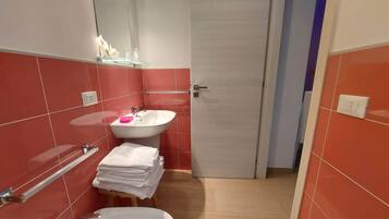 Un baño pequeño con un lavamanos blanco, un espejo y un inodoro. Hay un perchero con toallas dobladas y un estante de vidrio con artículos de aseo.