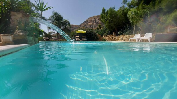 A clear blue pool with a waterfall feature, surrounded by lounge chairs and a stone wall. - La Suite di Segesta (Calatafimi)