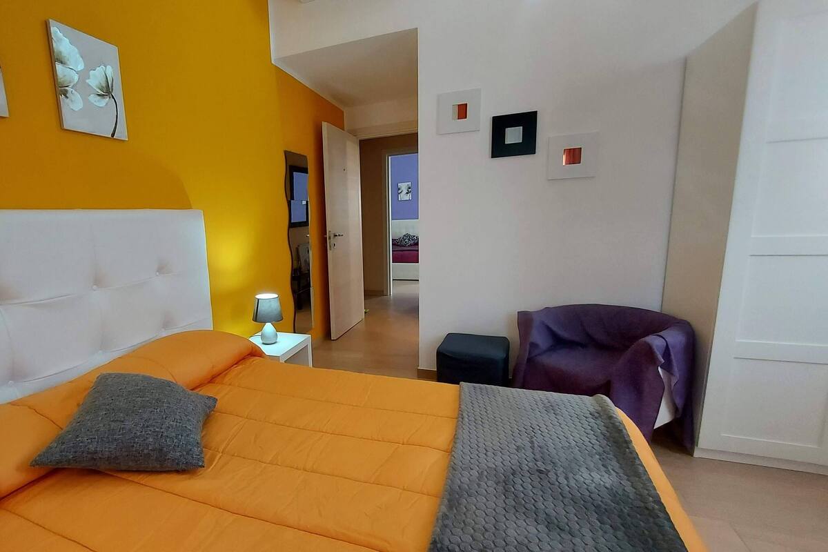 Quarto com cama grande, mesa de cabeceira, luminária, cadeira e obra de arte na parede.