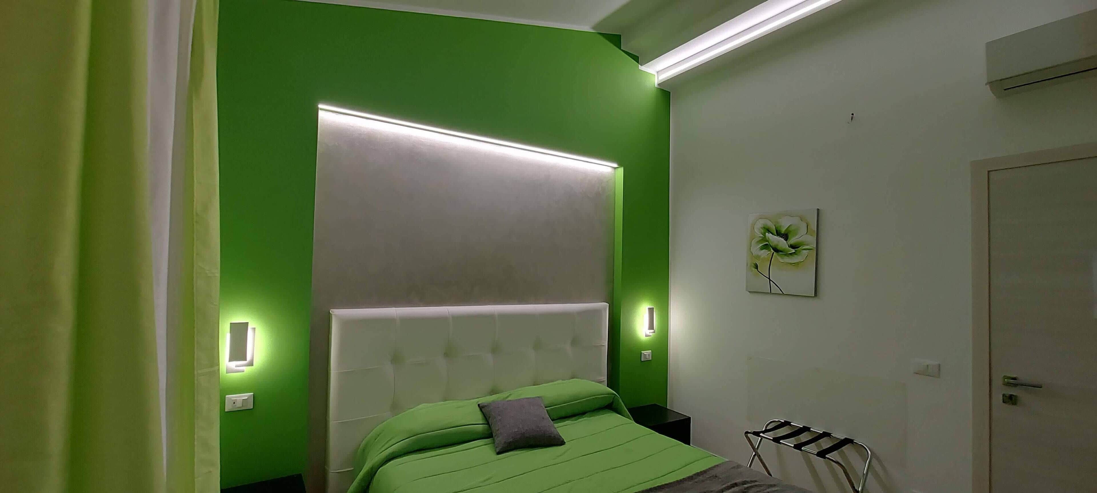 Un dormitorio con pared verde, cabecera blanca y cama con ropa de cama verde. Hay una mesita de noche con una lámpara, una luz de pared y un cuadro.