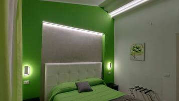 Un dormitorio con pared verde, cabecera blanca y cama con ropa de cama verde. Hay una mesita de noche con una lámpara, una luz de pared y un cuadro.