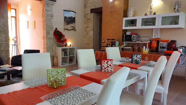 A dining area with white chairs and tables, a stone wall, and a kitchen area with appliances. - La Suite di Segesta (Calatafimi)