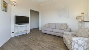 Uma sala de estar com uma TV de tela plana sobre um console branco, um sofá bege e uma poltrona estampada.