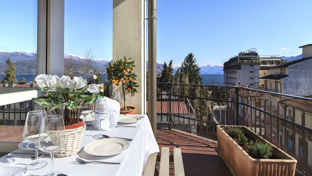 Varanda com mesa posta para refeições, com vista para uma paisagem montanhosa e um corpo d'água.