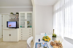 A modern living room with a television, a dining table with a magazine, a bowl of fruit, and a bottle of wine.