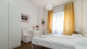 Habitación de hotel con dos camas individuales, un armario blanco, una mesita de noche pequeña con lámpara y una ventana con cortinas amarillas.