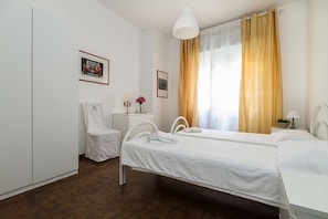 A hotel room with two single beds, a white wardrobe, a small bedside table with a lamp, and a window with yellow curtains. - Impero House Rent - Cavour (Stresa)
