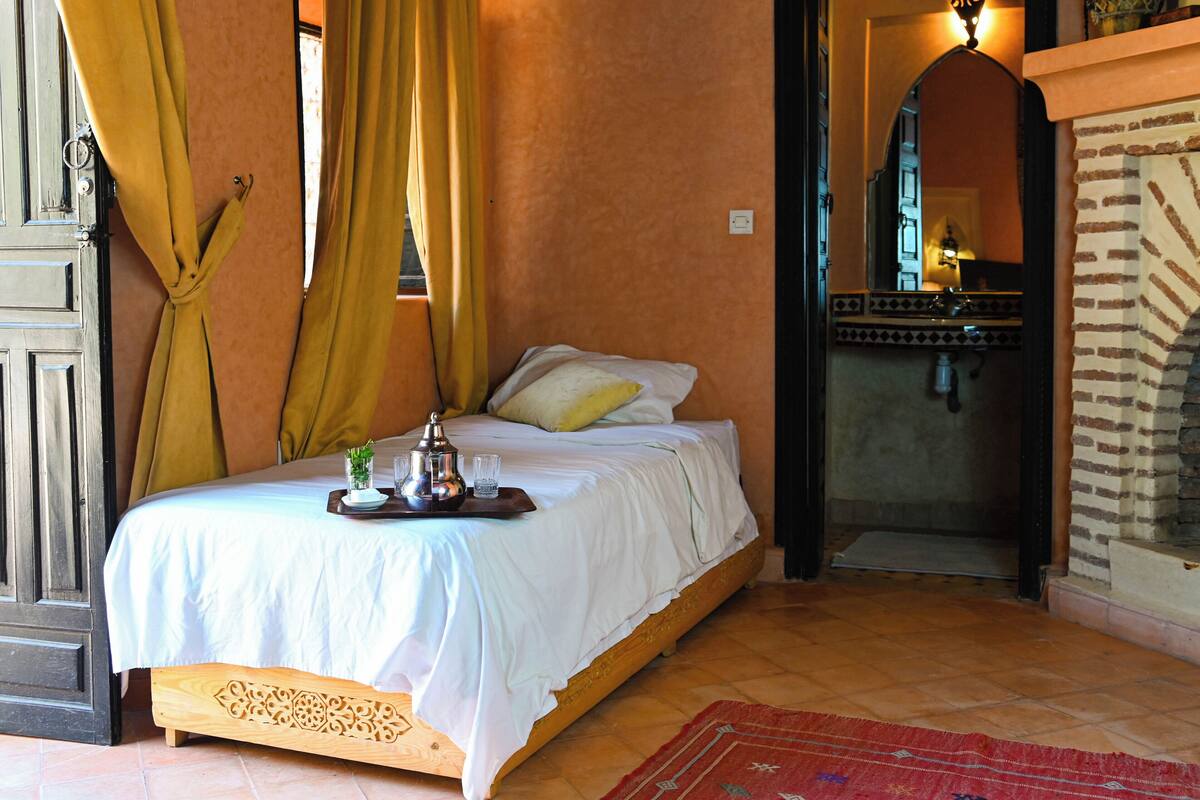 A bedroom with a bed, curtains, a small table with a tray, and a view of a kitchen area.