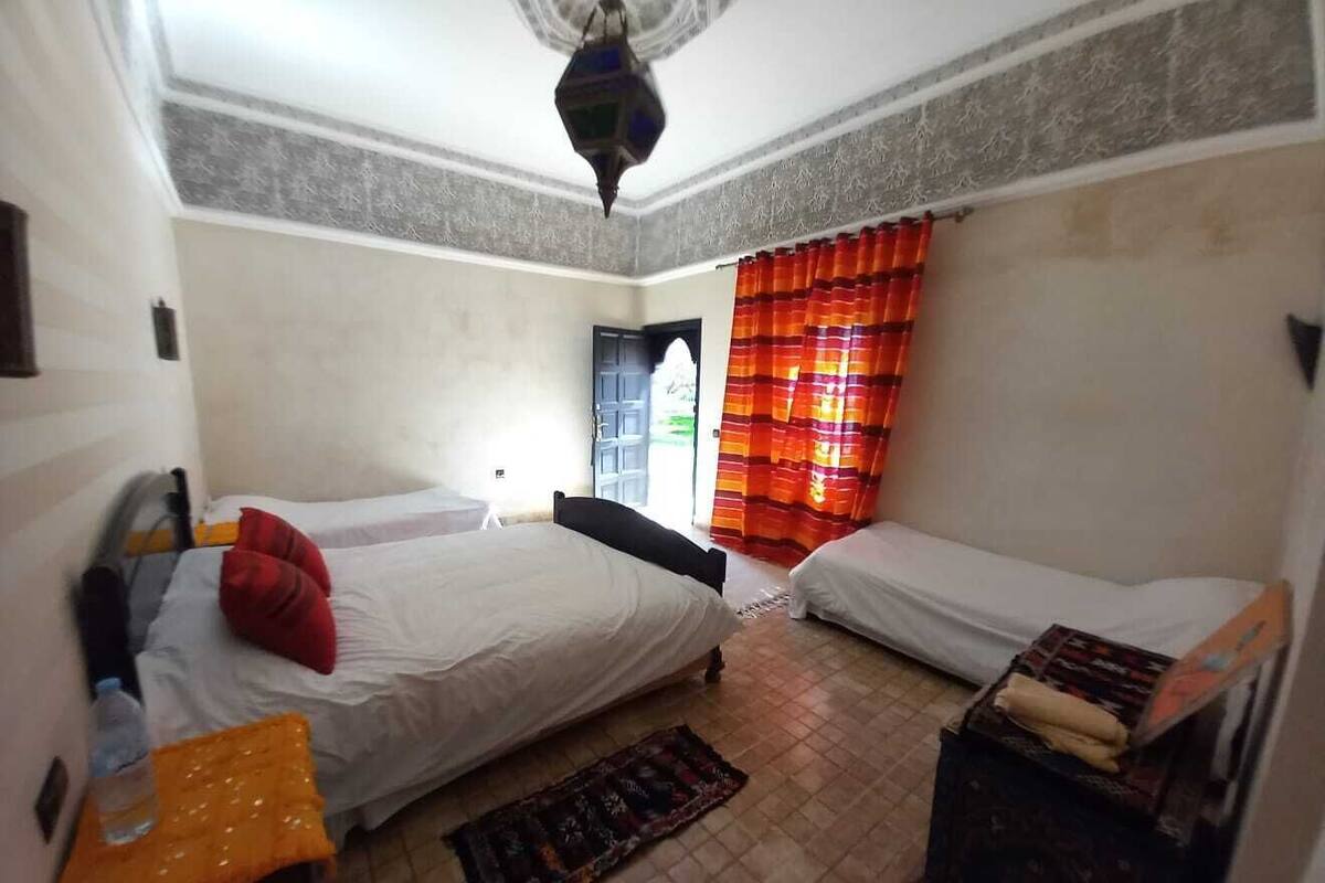 A room with two beds, a ceiling fan, a window with red curtains, and a small table with a box on it.