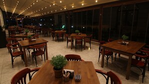Un restaurante espacioso con múltiples mesas y sillas de madera, plantas en macetas y vistas amplias a través de grandes ventanales.