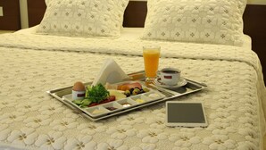 Una cama bien tendida con una bandeja de desayuno, que incluye tostadas, fruta y un vaso de zumo de naranja, sobre una cama de hotel con una colcha de estampado floral y dos almohadas.