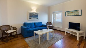 Uma sala de estar com um sofá azul, uma mesa de centro branca, uma cadeira de madeira, uma televisão em um móvel e um quadro na parede.