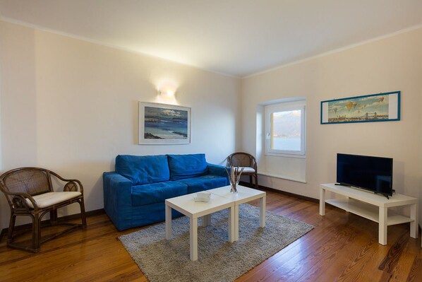 A living room with a blue sofa, a white coffee table, a wooden chair, a TV on a stand, and a painting on the wall.
