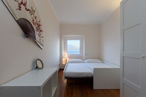 A small, well-lit room with a single bed, a bedside table, a decorative fan, and a window with a view. - Impero House Rent - Bella Vista (Stresa)