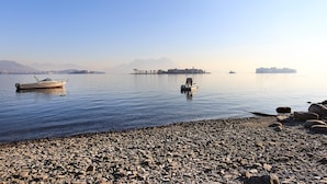 A calm lake with boats, a rocky shore, and distant buildings. - Impero House Rent - Maison du Lac (Baveno)
