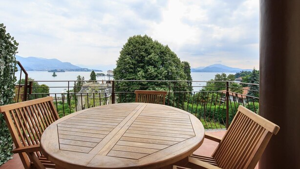 A round wooden table with four chairs on a balcony overlooking a lake with mountains in the background. - Impero House Rent - Rampolina View (Stresa)