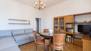 A modern living room with a grey sofa, wooden dining table, and chairs, a chandelier, a television, and a bookshelf. - Impero House Rent - Rampolina View (Stresa)