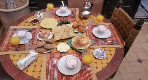 Una mesa de comedor redonda con diversos alimentos para el desayuno, como pan, huevos y bollería, sobre un mantel estampado.
