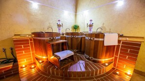 A sauna with wooden benches and a central bench with a towel. There are two sinks with taps and a plant in the background. - LH Hotel Dvorak Tabor (Tabor)