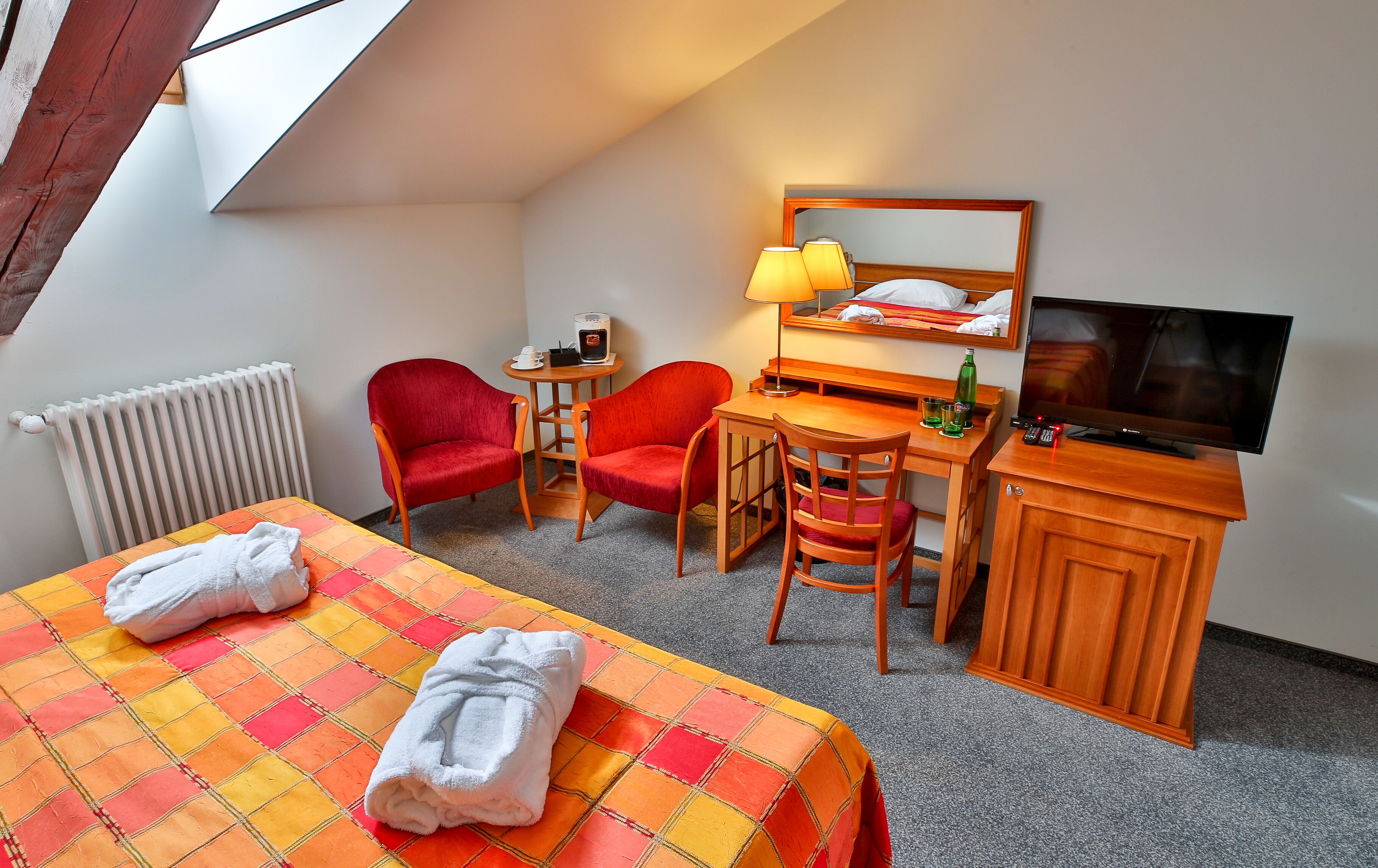 A hotel room with a bed, two red chairs, a desk with a lamp, a television, and a radiator.