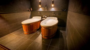 A modern bathroom with two wooden tubs, dark tiled walls, and wall-mounted lighting.
