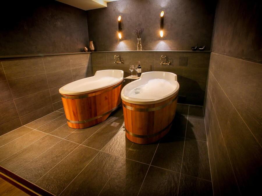 A modern bathroom with two wooden tubs, dark tiled walls, and wall-mounted lighting.