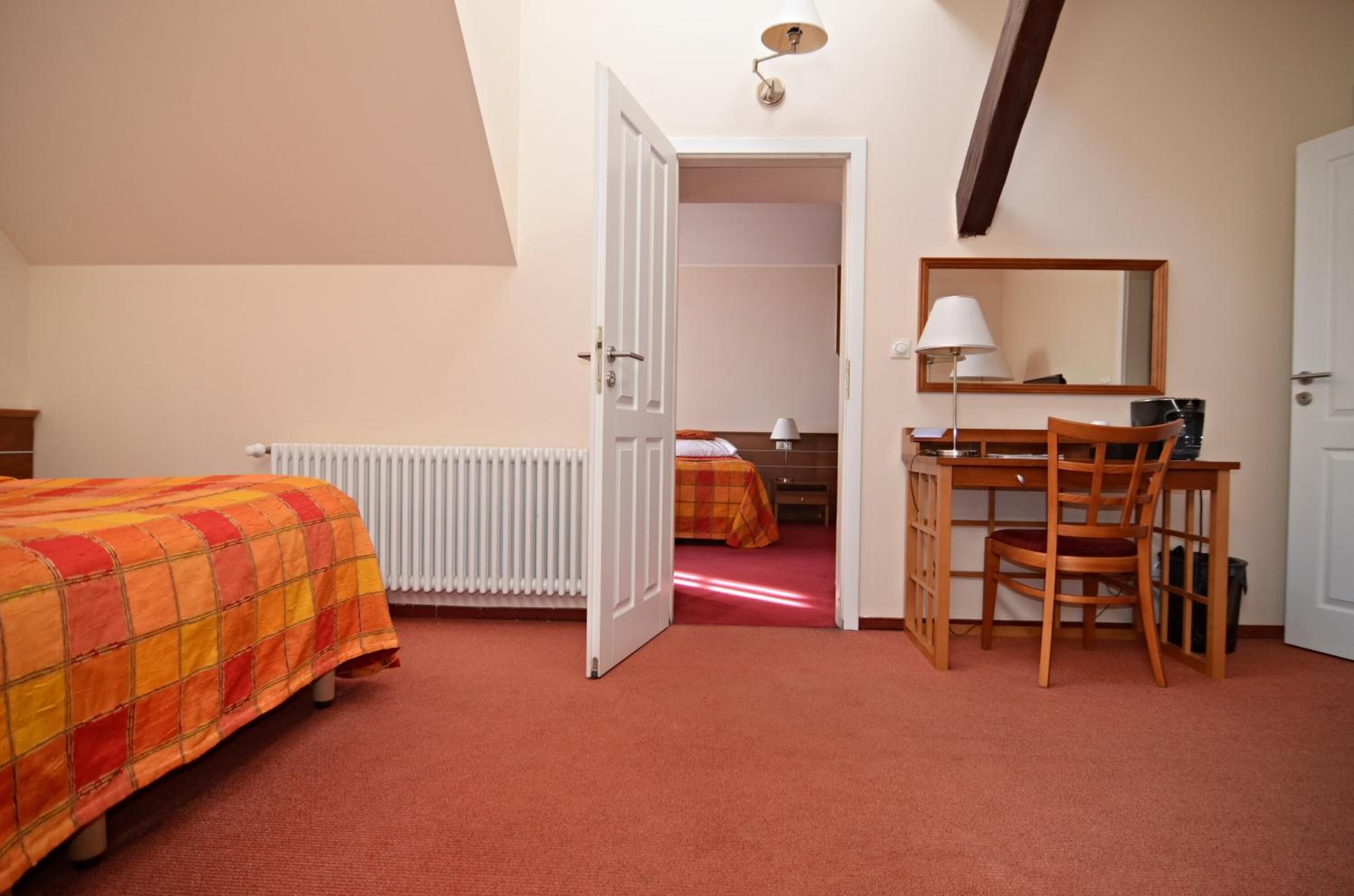 A bedroom with a bed, a desk, a chair, a radiator, a mirror, and a door leading to another room.