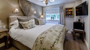 A bedroom with a large bed, a heart-shaped wall decoration, a chandelier, a window with curtains, a shelf with jars, and a television. - The Grousemoor Country House (Wrexham)