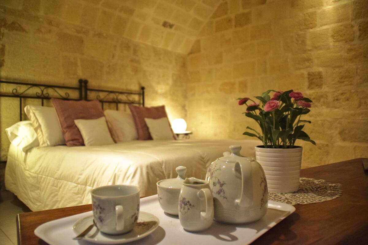 Un lit bien fait, agrémenté d’oreillers, une table de chevet sur laquelle est posé un plateau avec un service à thé, et une plante en pot aux fleurs roses.
