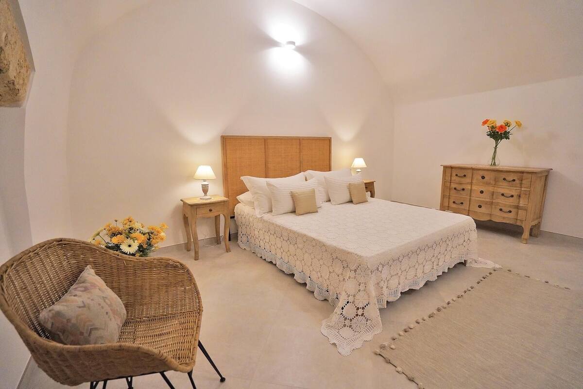 Une chambre avec un grand lit, une chaise en osier, une table de chevet avec une lampe, une commode et un vase rempli de fleurs.