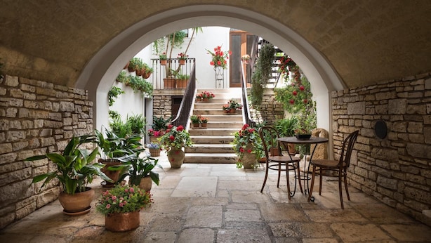 Un corredor de piedra con arcos, macetas con plantas y una escalera que lleva a una puerta de madera.