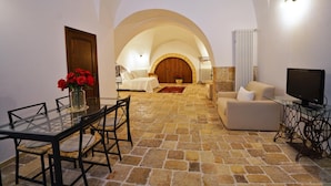 Un salón acogedor con suelo de piedra, una mesa de vidrio para comer con sillas, un sofá, un televisor sobre un atril metálico y una puerta de madera que da a otra habitación.