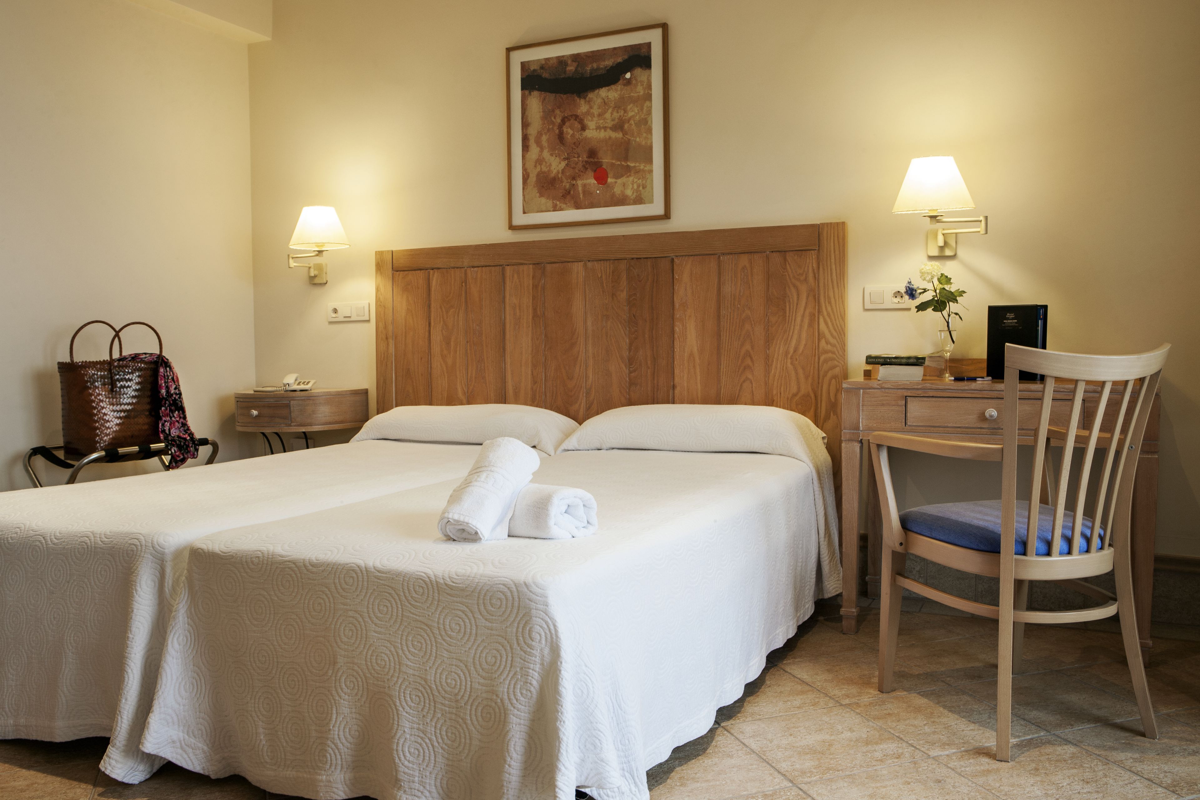 Camera d'albergo con due letti, una testiera in legno, una scrivania con sedia, una lampada e un quadro appeso al muro.