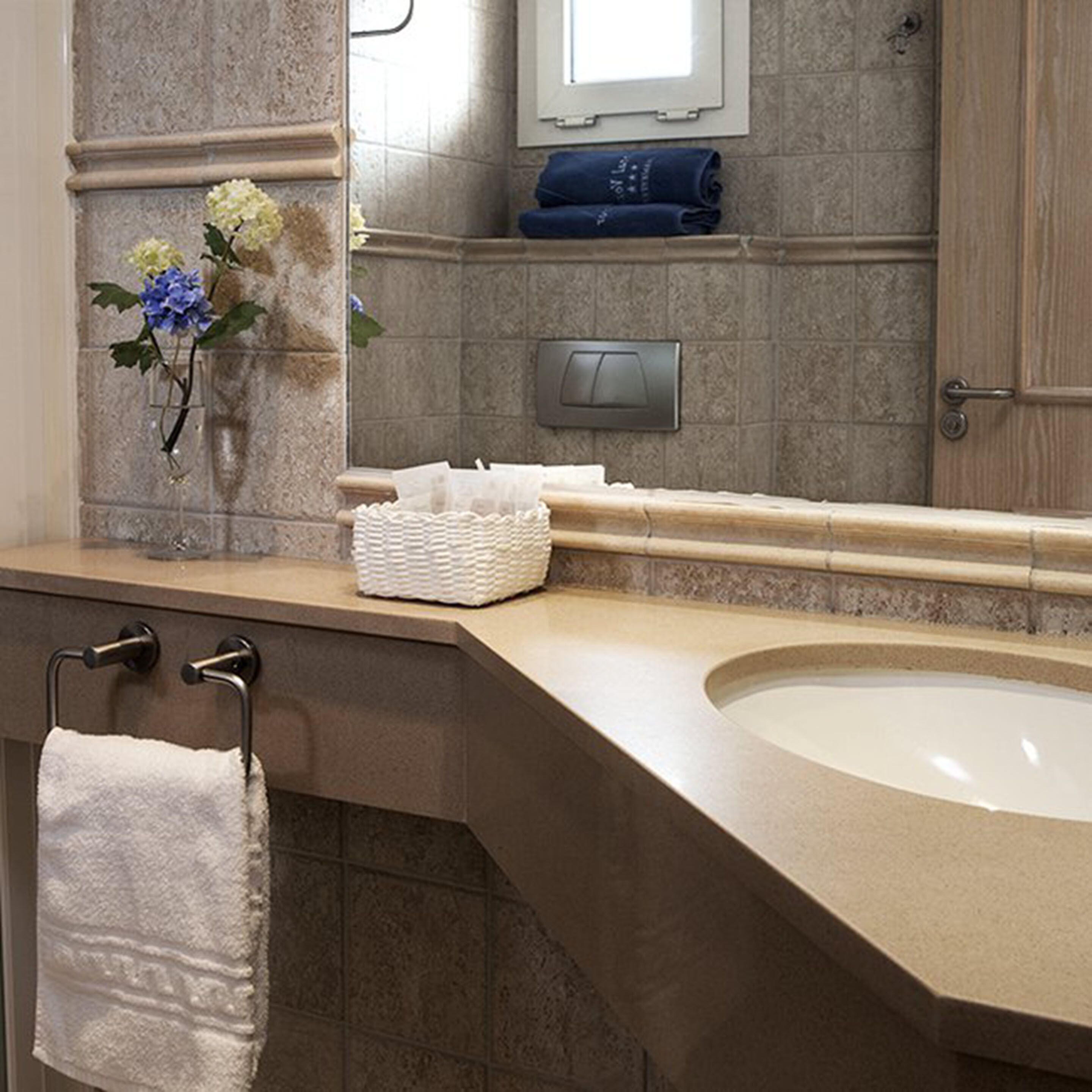 Een badkamer met een beige wastafel, een handdoekhouder, een witte mand en een vaas met bloemen.