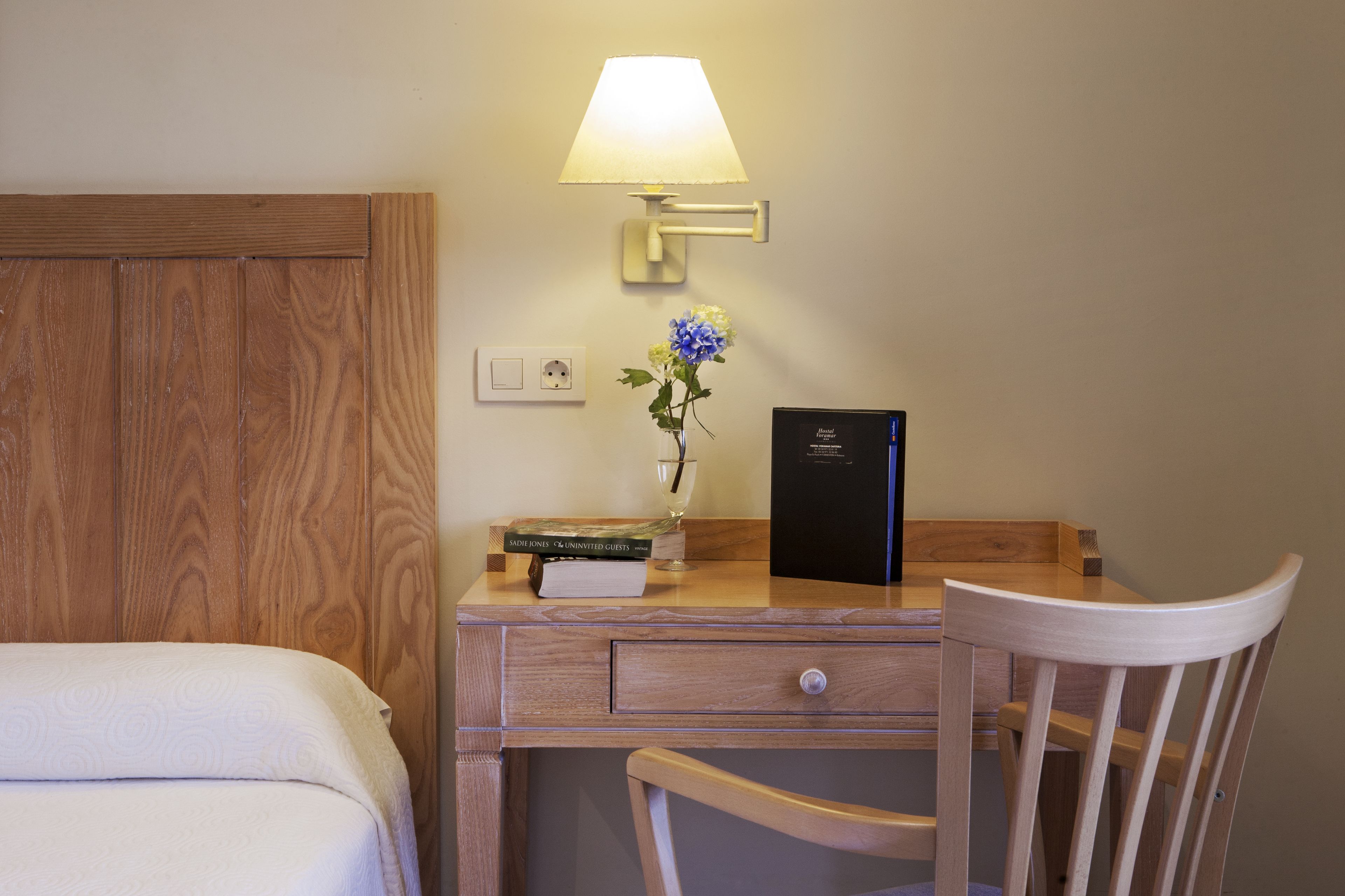 A wooden desk with a lamp, a vase with flowers, a book, and a black box, likely a minibar, in a hotel room.