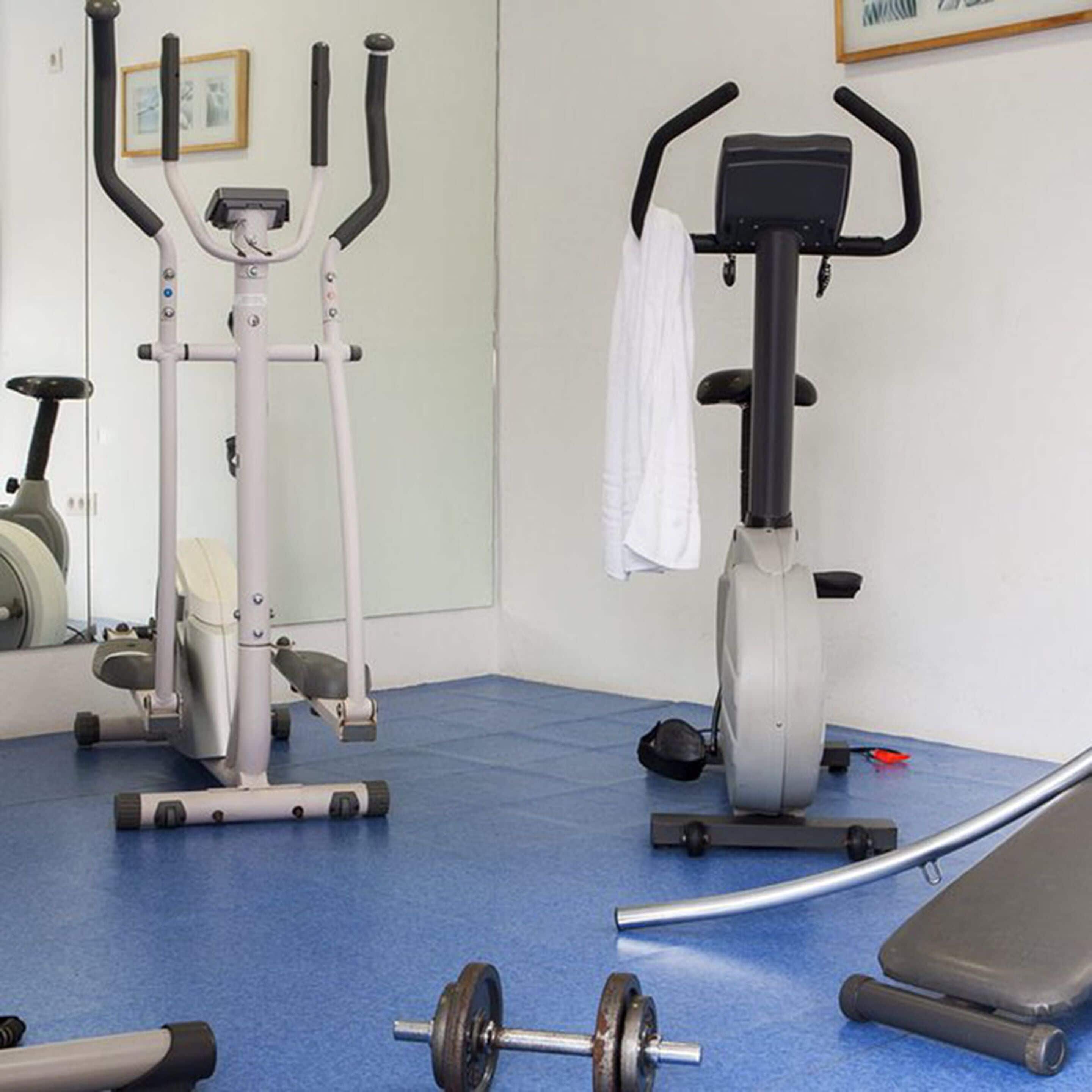 Ein Fitnessraum mit Trainingsgeräten, darunter ein Ergometer, ein Crosstrainer und eine Hantel auf dem Boden.