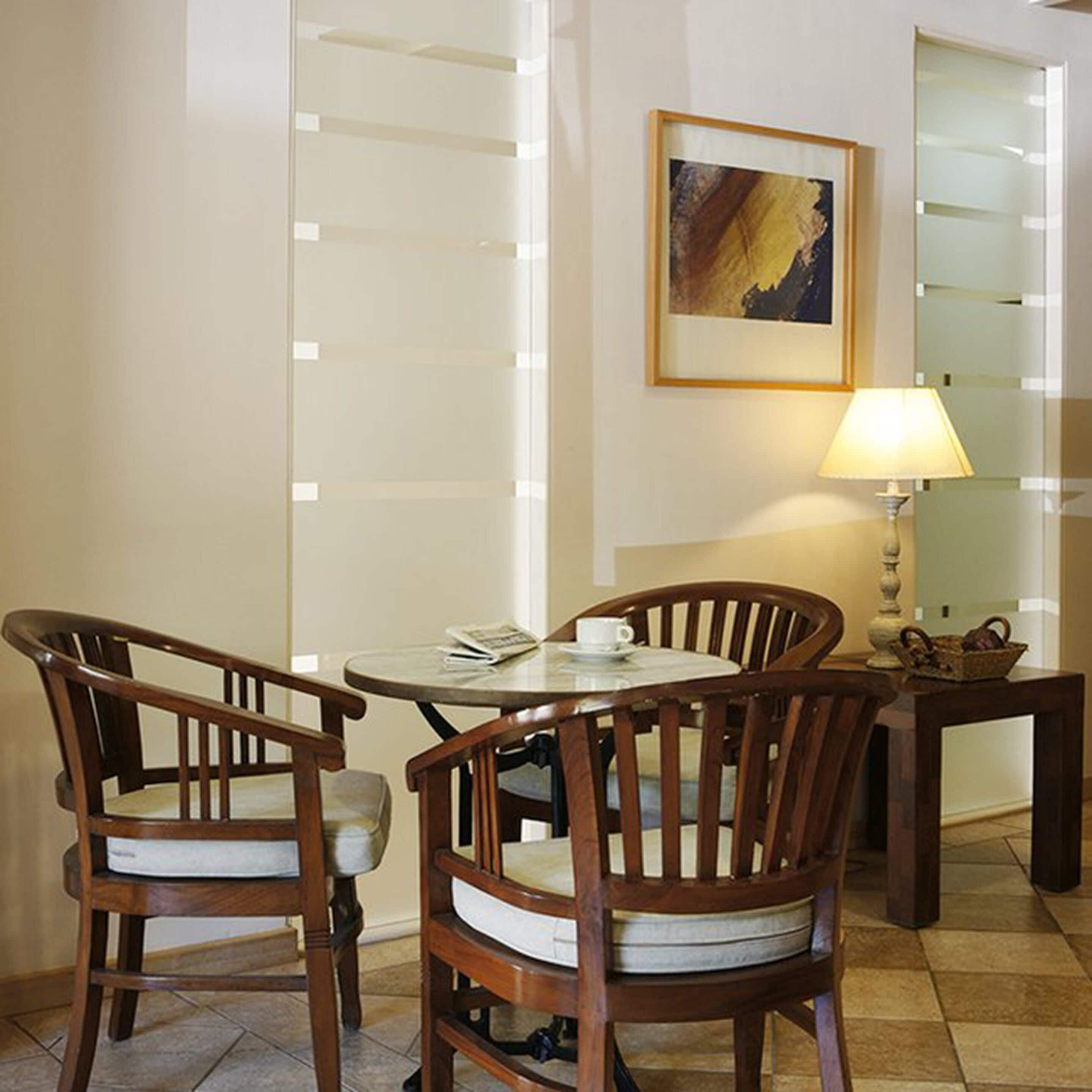 A dining area with two wooden chairs, a small round table, a lamp, and a framed painting on the wall.