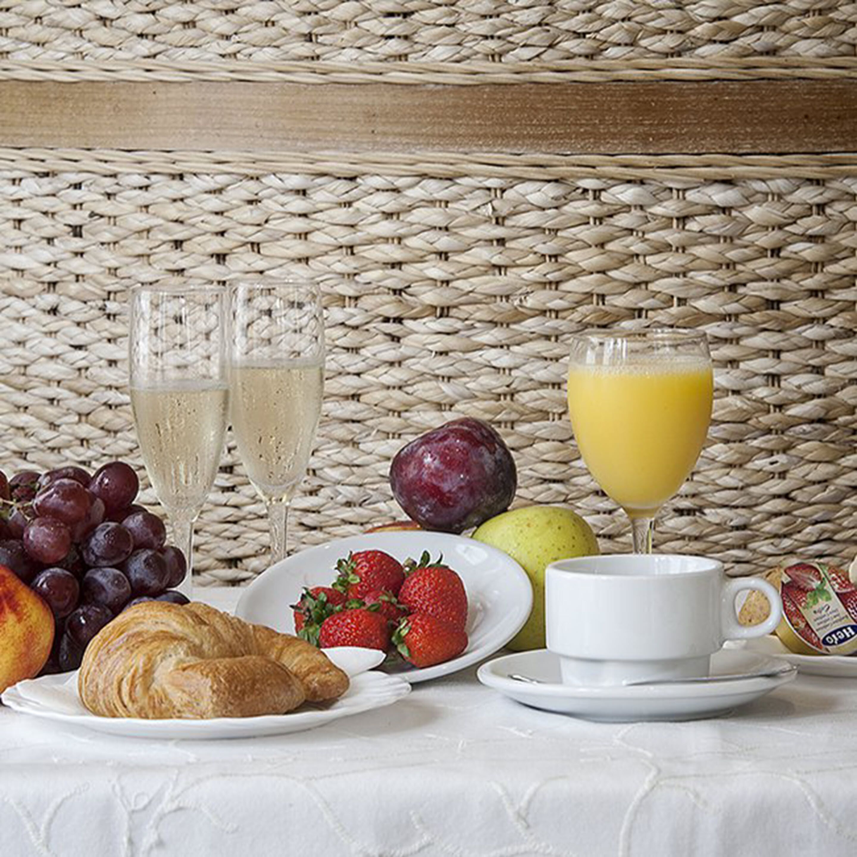 A breakfast setting with a woven tablecloth, two glasses of white beverage, a plate of strawberries, a croissant, a glass of orange juice, and a bowl of yogurt.
