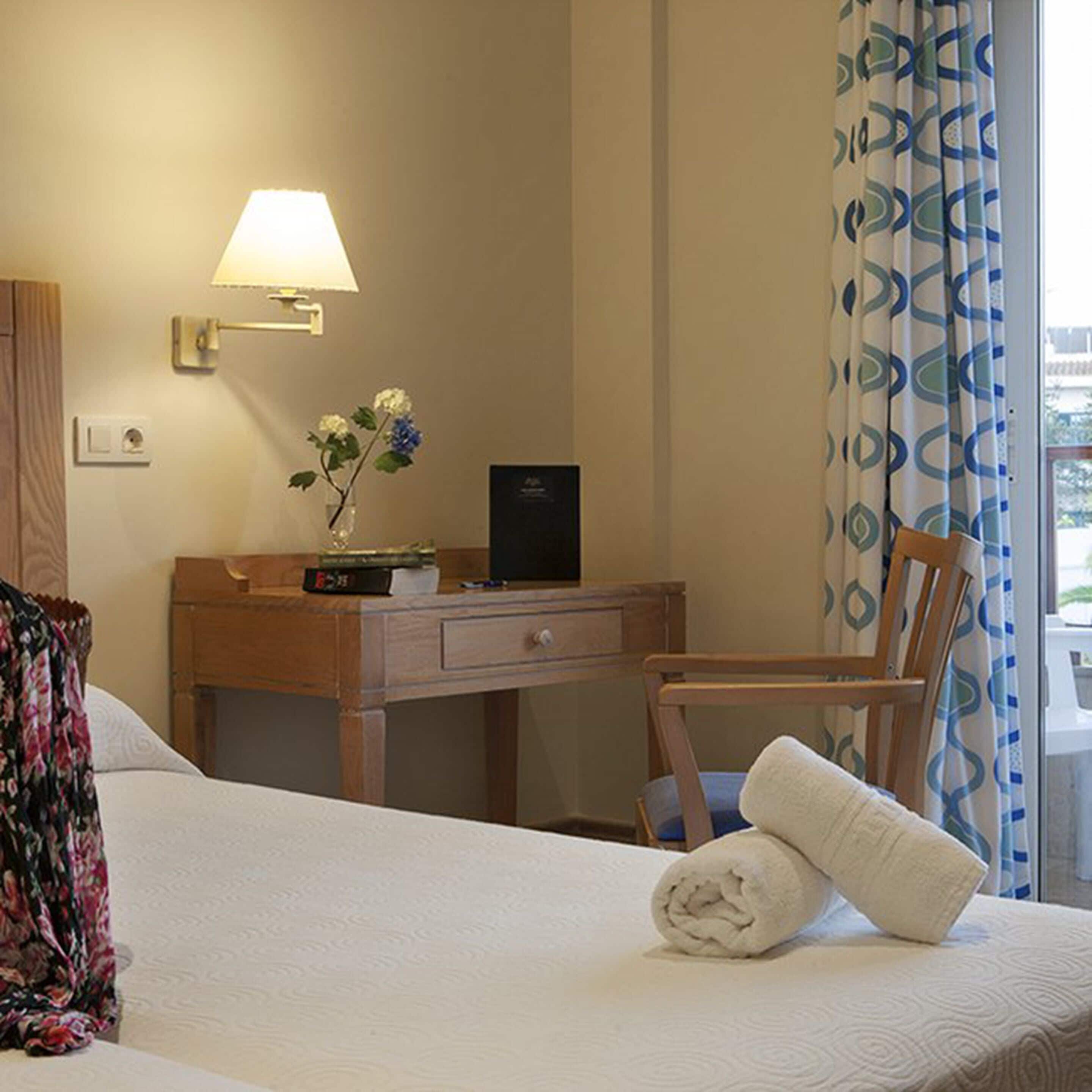 Een hotelkamer met een bed, een bureau met een lamp, een stoel, een tafeltje met een bloevas en een raam met gordijnen.