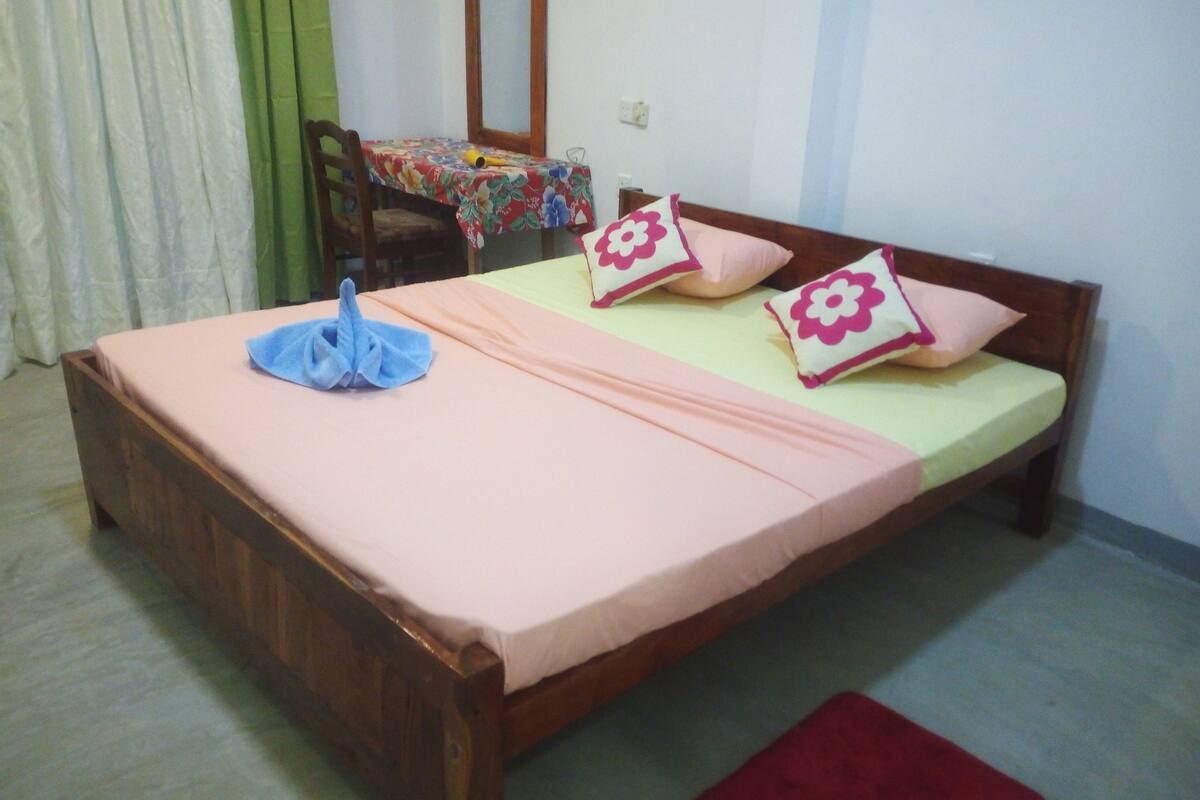 A single bed with a wooden frame, a pink bedspread, and two decorative pillows with floral patterns.