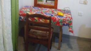 Una habitación pequeña con una sola silla de madera, una mesa con mantel floreado, un espejo en la pared y una cortina verde.