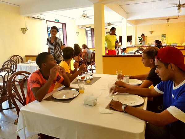 A dining area with tables, chairs, and people eating. There are signs indicating an exit and a water dispenser. - Ayati Hotels (Flic-en-Flac)