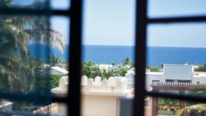 A view through a window of a coastal area with buildings, palm trees, and the ocean. - Ayati Hotels (Flic-en-Flac)