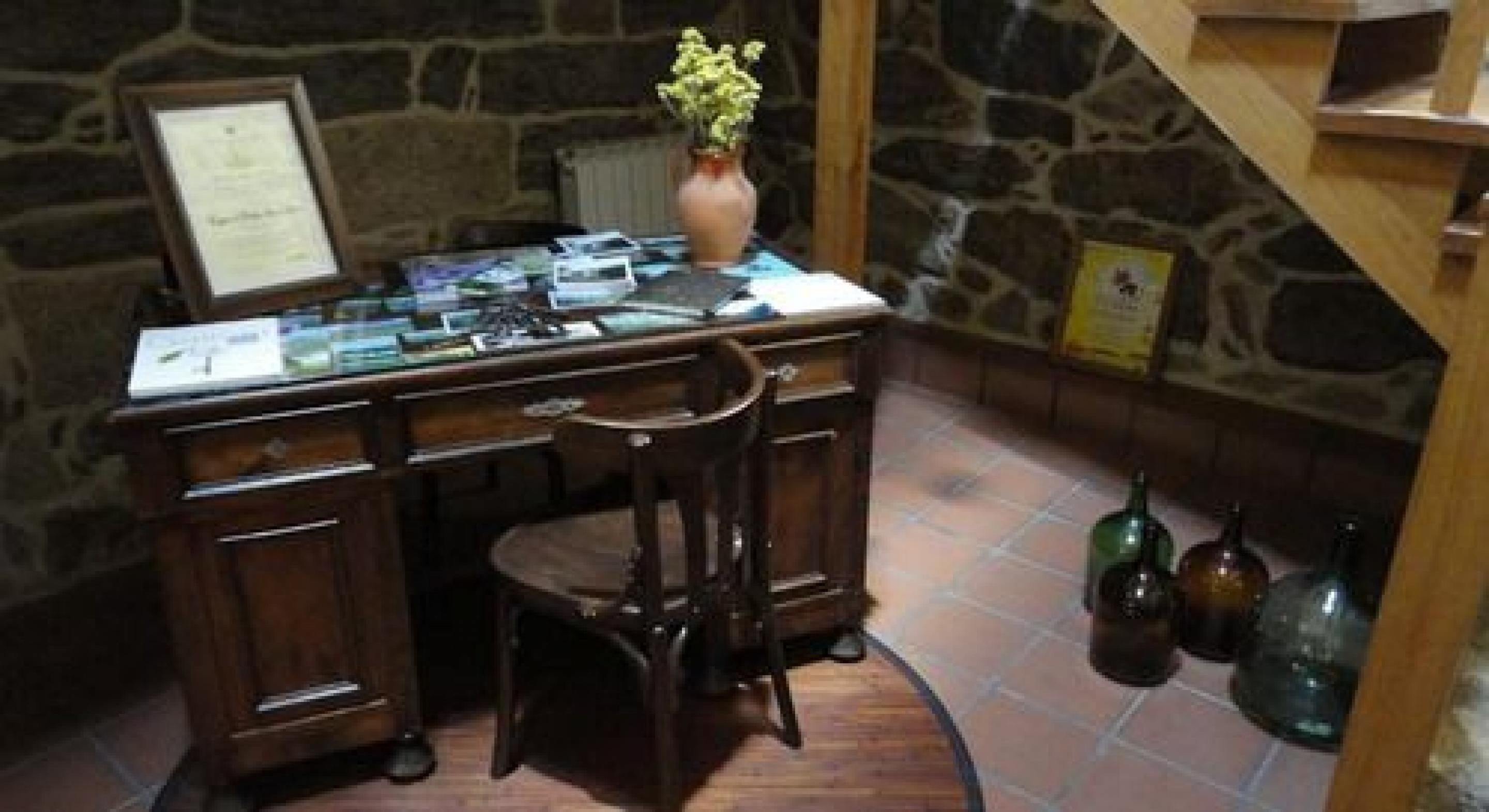 A wooden desk with a vase of flowers, a framed certificate, and a computer. There are two bottles on the floor.