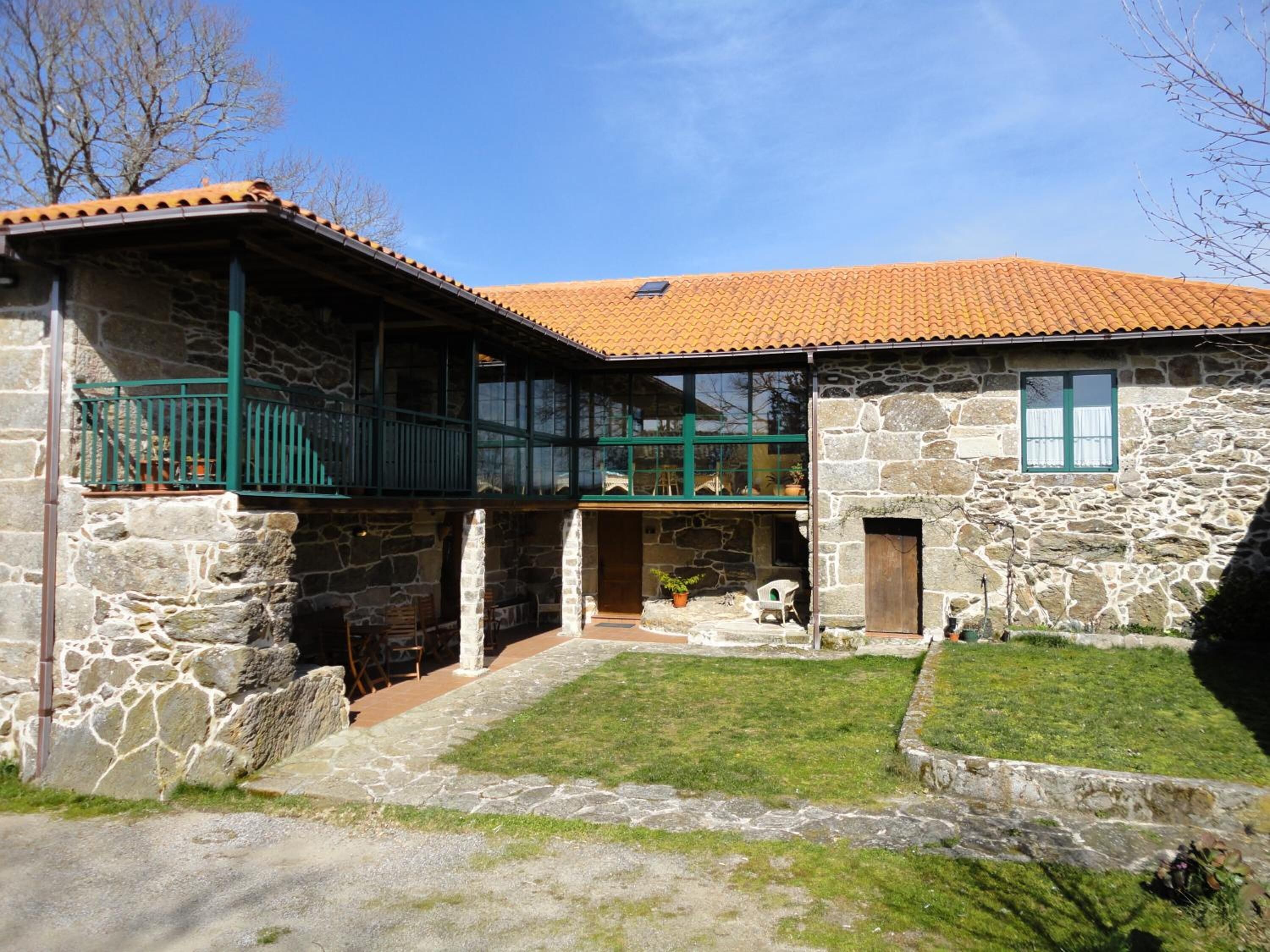 A stone house with a tiled roof, a balcony with green railings, and a small garden area.