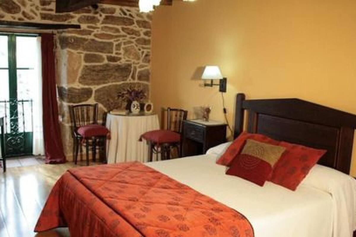 A bedroom with a stone wall, a bed with red and white bedding, a desk with a lamp, and a chair.