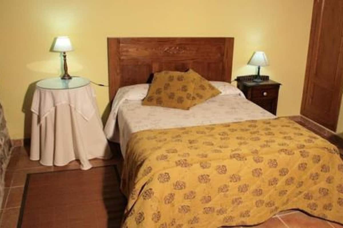 A bedroom with a wooden headboard, a bed with a patterned bedspread, two bedside tables with lamps, a chair, and a small table with a tablecloth.