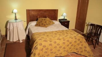 A bedroom with a wooden headboard, a bed with a patterned bedspread, two bedside tables with lamps, a chair, and a small table with a tablecloth.